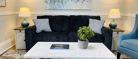 Una sala de estar con un sofá azul oscuro, dos lámparas a juego, una mesa de centro blanca y un cuadro abstracto grande en la pared.
