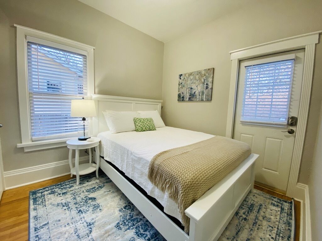 Un dormitorio con una cama blanca, una mesita de noche pequeña con una lámpara, una alfombra azul y blanca estampada y una puerta con un ventanal.