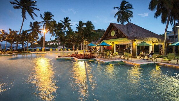 Área de piscina en un resort con un edificio de techo de paja, palmeras y vista al océano.