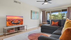 A modern living room with a flat-screen TV, a grey sofa with orange cushions, a patterned rug, and a large window with curtains. - Modern and Sophisticated Old Town Scottsdale Gem! (Scottsdale)
