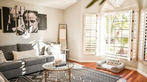 Un salon avec un canapé gris, une table basse en verre et un tapis à motifs. Il y a une fenêtre avec des stores et une vue sur la verdure à l’extérieur.
