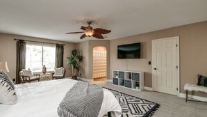 Une chambre spacieuse avec un grand lit, un ventilateur de plafond, une télévision, une bibliothèque et une porte.