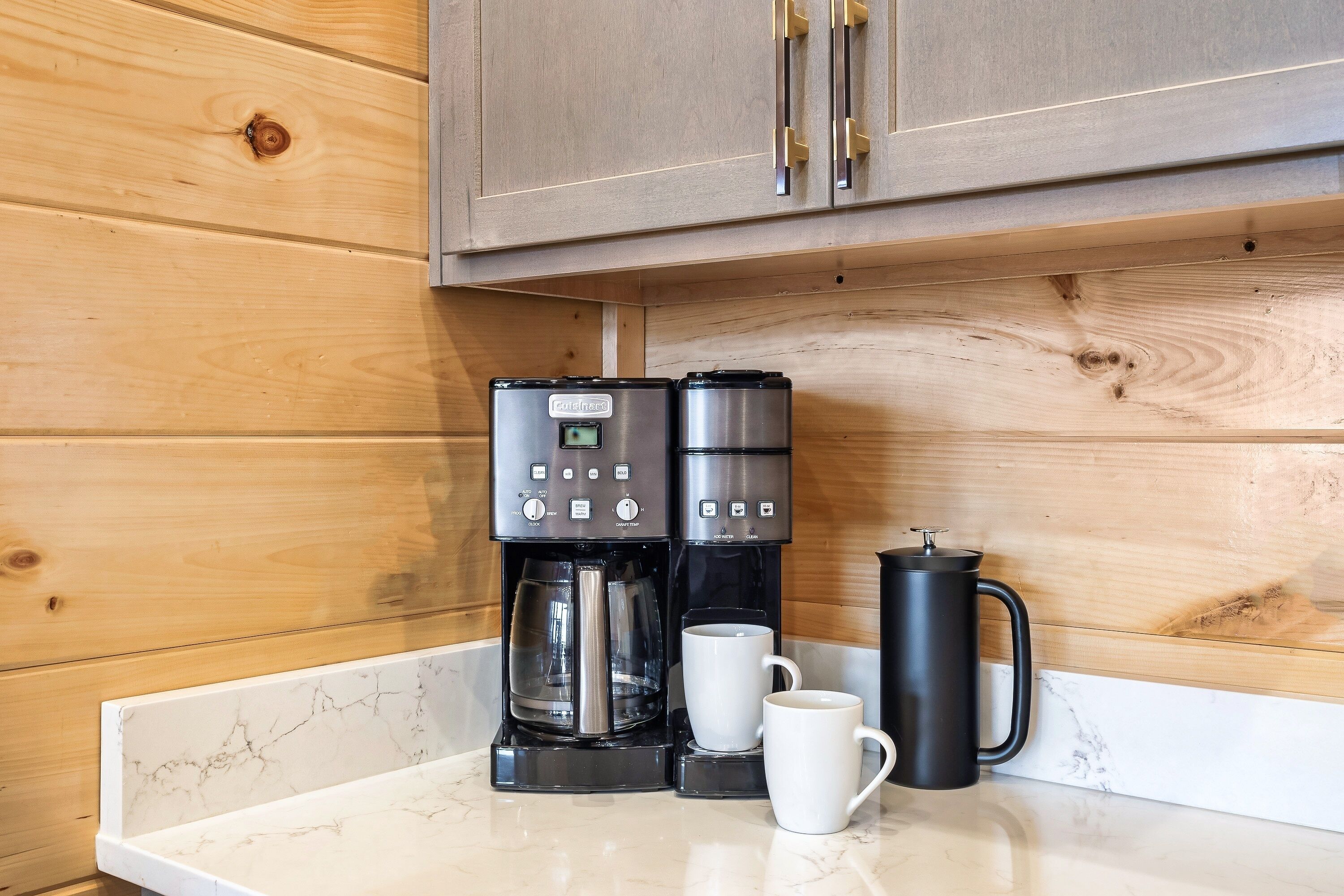 Eine Küche mit Kaffeemaschine, zwei weißen Tassen und einem schwarzen Thermosbehälter auf einer Marmorarbeitsplatte.