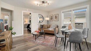 A modern living room with a sofa, coffee table, and dining area. There is a ceiling light fixture and a door leading to another room. - Charming Old Town Bungalow W/ Free Cruiser Bikes (Fort Collins)