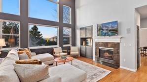 A spacious living room with a large gray sofa, a fireplace, and a television mounted above it. - Spacious Home W/ Fun Amenities | Lake & Mtn Views (Loveland)