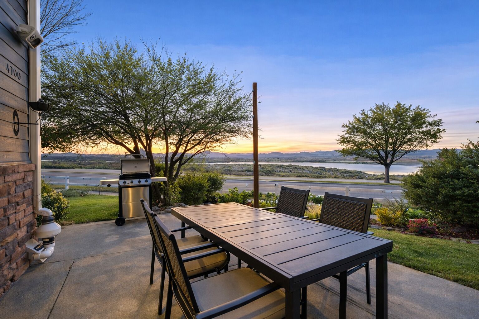 An outdoor patio with a grill, table, and chairs, overlooking a scenic landscape with a body of water and distant mountains.