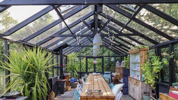 Un invernadero con una mesa y sillas de madera, rodeado de plantas en macetas y un mueble de madera con estantes.