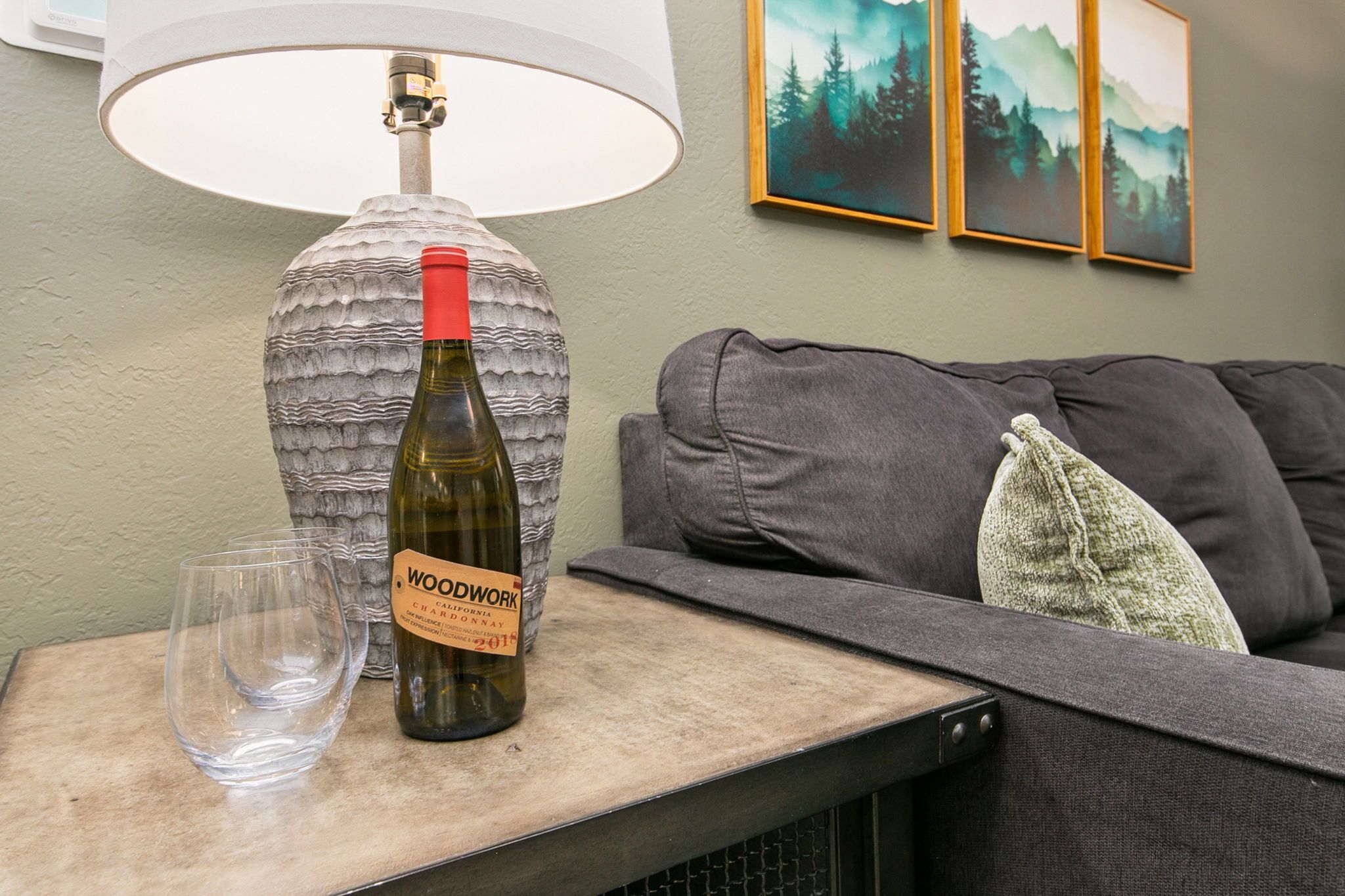 A wine bottle on a table with a lamp and a glass, a grey couch, and framed artwork on the wall.