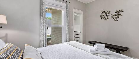 Un dormitorio con cama, una mesita de noche con toalla, una lámpara y una ventana con cortinas.