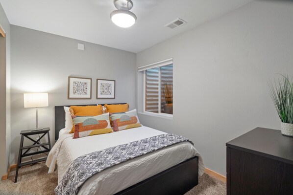 A bedroom with a bed, bedside lamp, two framed pictures, a window with blinds, and a small plant on a dresser. - Sleek Home Near Old Town W/ Rftp Hot Tub & Mtn Views (Fort Collins)