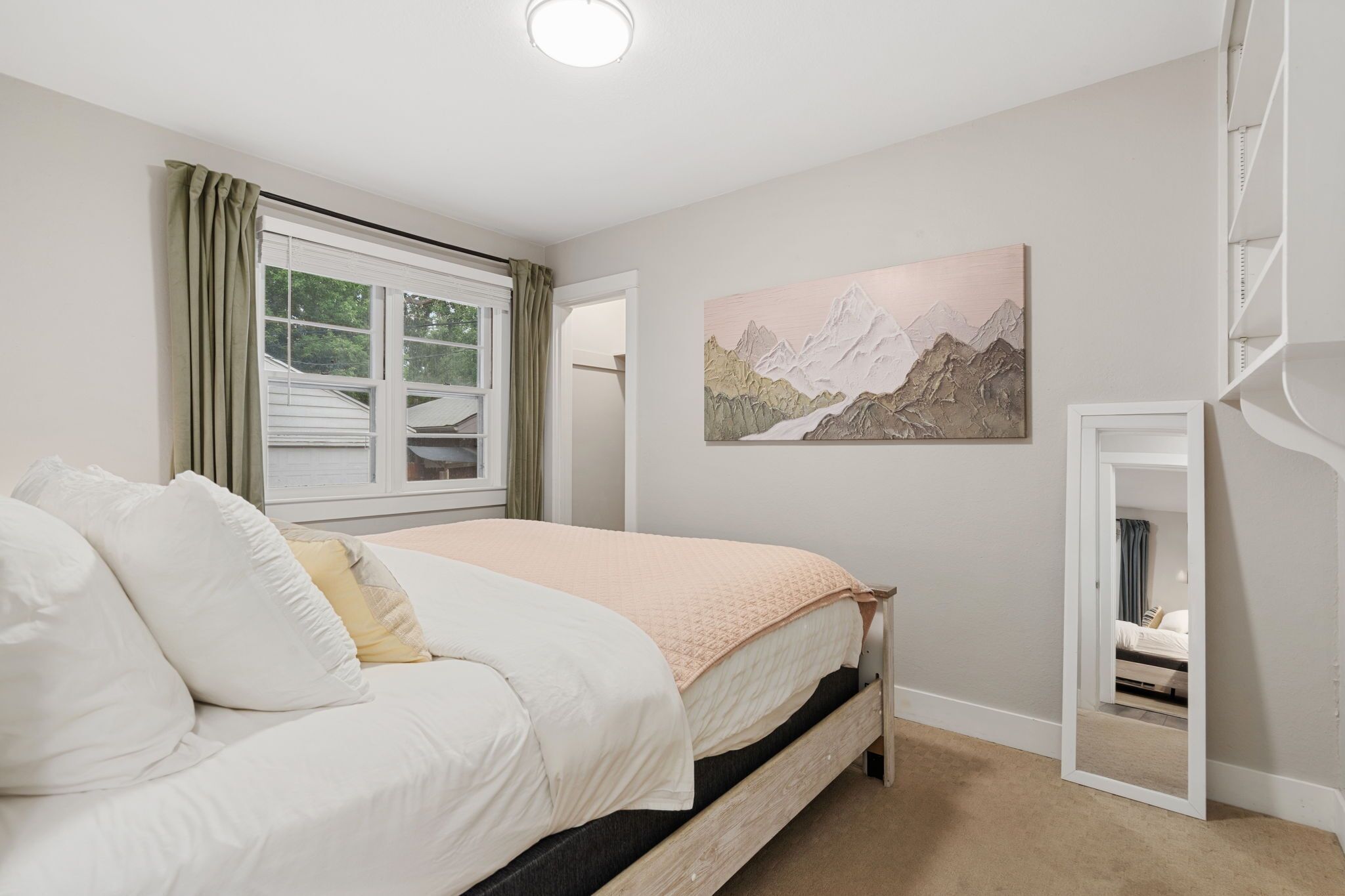 A bedroom with a bed, a window with curtains, a painting on the wall, and a mirror.