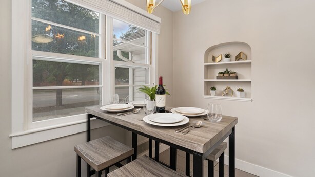A dining area with a wooden table set for four, a wine bottle, and a view of the street through large windows. - Beautiful Bungalow Next to Csu - Free Cruiser Bikes (Fort Collins)
