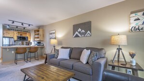 Un salon moderne avec un canapé gris, une table basse en bois et un coin cuisine à l’arrière-plan.