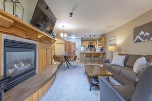 Cozy living area with a fireplace, comfortable sofa, and dining table for four, complemented by a kitchen area with modern appliances and a bar.