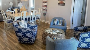 A spacious living room with a wooden floor, a dining area with a white table and chairs, and a kitchen in the background. - 4 BR, 3 bath Cherry Grove Retreat near CG Pier, Private Pool, 1 block to beach (North Myrtle Beach)