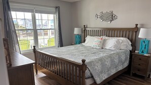 A bedroom with a wooden bed, two bedside tables with lamps, a window with curtains, and a wall decoration. - 4 BR, 3 bath Cherry Grove Retreat near CG Pier, Private Pool, 1 block to beach (North Myrtle Beach)