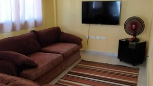 A living room with a large brown sofa set, a flat-screen TV mounted on the wall, and a standing fan on a small table. - CASA DE PRAIA NA LINDA MARAGOGI (Maragogi)