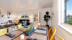 Een moderne woonkamer met een bank, eettafel, gele stoelen en een groot raam met uitzicht op groen.