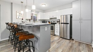 A modern kitchen with white cabinets, stainless steel appliances, a central island with bar stools, pendant lighting, and a view of the outdoors.