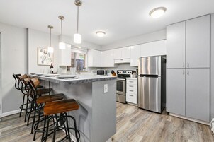 Cocina moderna con armarios blancos, electrodomésticos de acero inoxidable, una isla central con taburetes y luminarias colgantes, además de vistas al exterior.