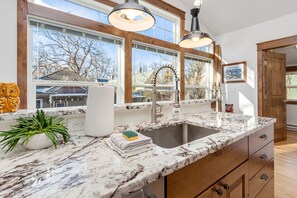 A kitchen with a marble countertop, wooden cabinets, a stainless steel sink, and a window with a view of a house and trees. - The Beautiful Bear’s Den! Walk to Old Town & CSU! (Fort Collins)