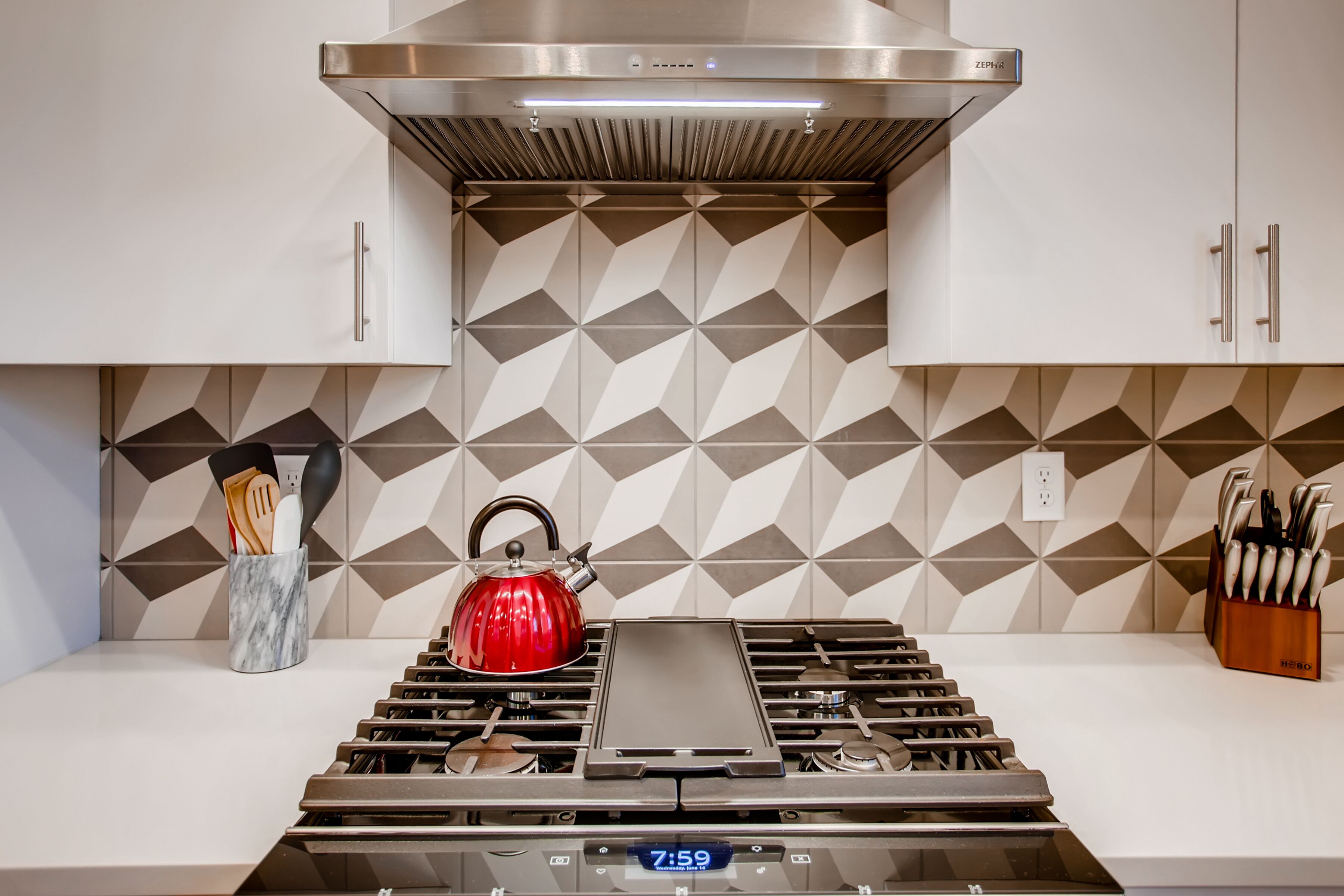 A kitchen with a modern stove, a red kettle, and a geometric backsplash.