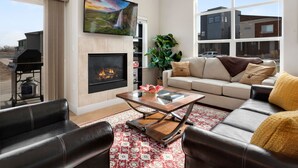 A living room with a fireplace, a sofa, a coffee table, and a large window offering a view of a house.