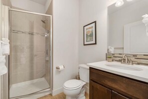 A bathroom with a glass shower enclosure, a toilet, a sink with a mirror, and a framed picture on the wall.