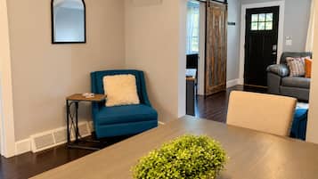 Una sala de estar moderna con un sillón azul, una mesa de comedor con un centro de mesa de planta verde y una puerta negra con ventana.