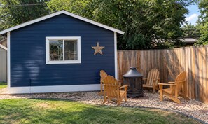 A blue shed with a window and a star decoration, surrounded by wooden chairs and a wood-burning stove.
