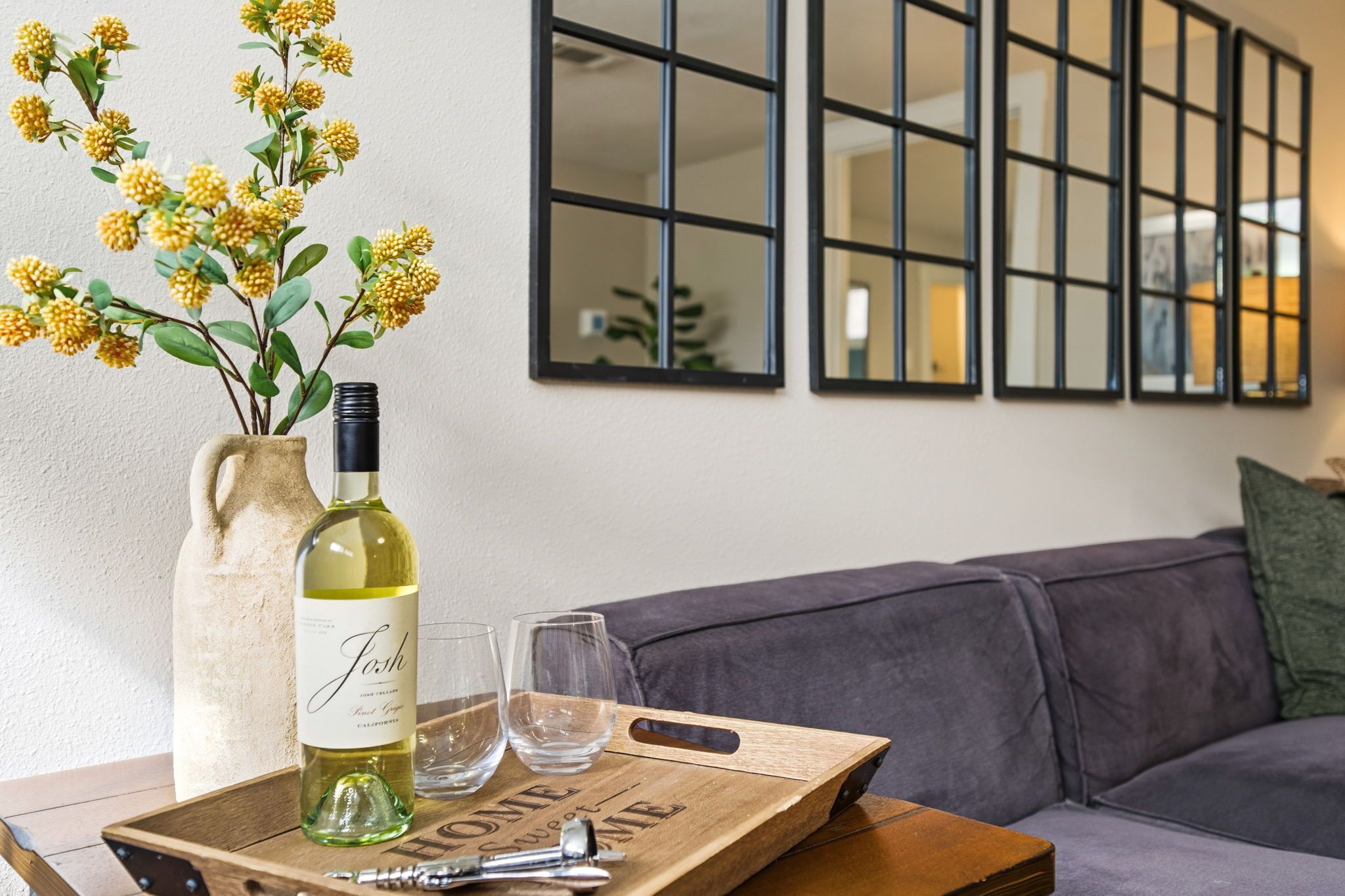 Una botella de vino, copas y una bandeja de madera sobre una mesa con un florero de flores amarillas.