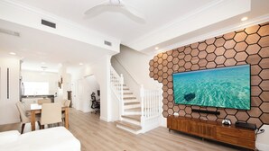 Een moderne woonkamer met een grote flatscreen televisie bevestigd aan een houten wand in een zeshoekig patroon, een trap, een eethoek en een keuken op de achtergrond.