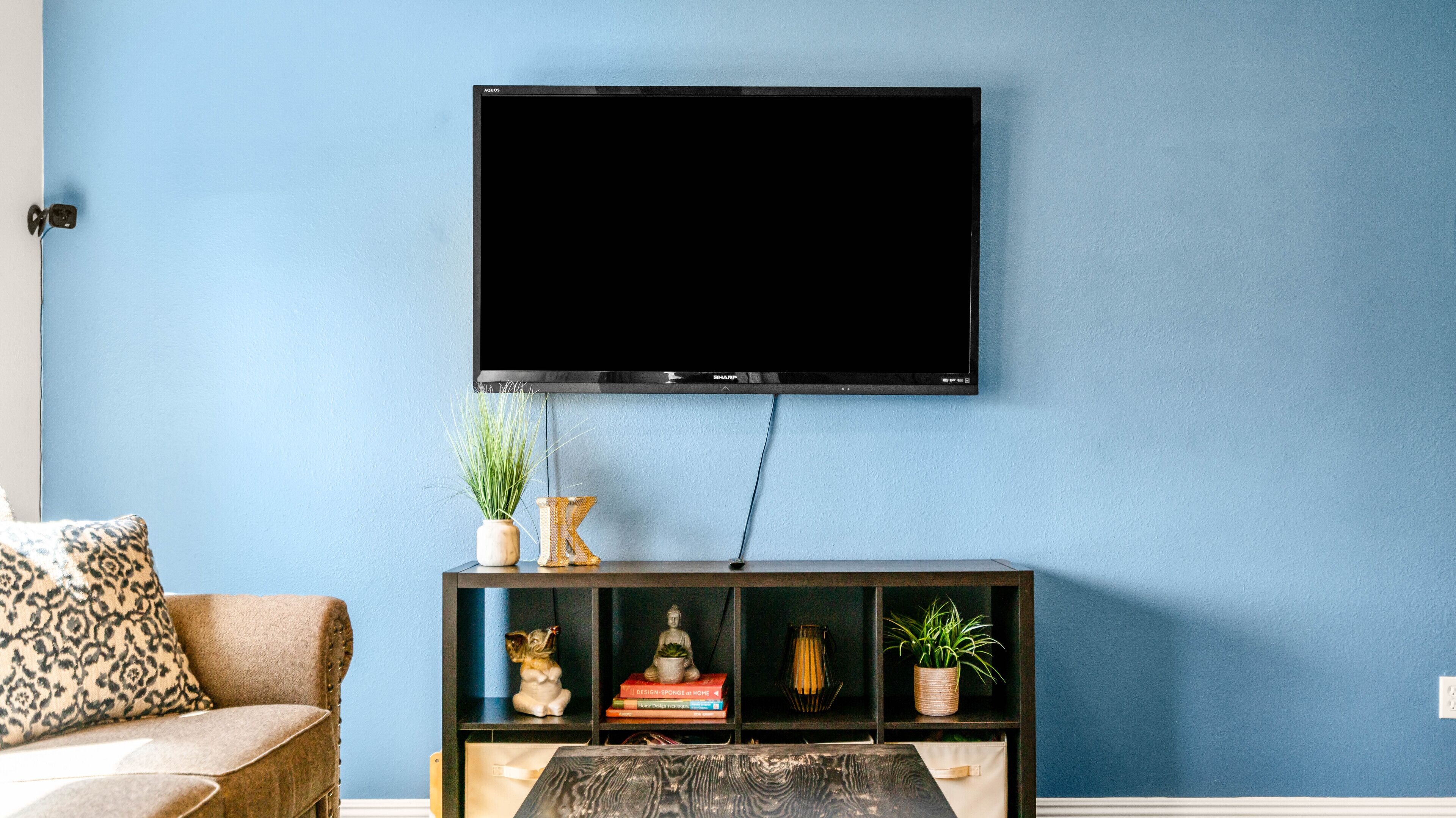 Un salon avec un téléviseur à écran plat fixé au mur, une console en bois foncé agrémentée d’objets décoratifs, un coussin à motifs sur un canapé et un mur bleu.
