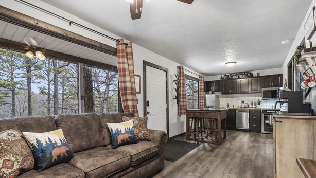 A cozy living room with a brown sofa and patterned cushions, a kitchen with dark wooden cabinets, and a large window overlooking trees. - Charming Rustic Retreat - Hot Tub - Pool - Coffee - Fire Pit - 1 mile from SDC! (Branson)