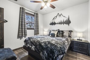 A bedroom with a bed, a nightstand, a window with curtains, a ceiling fan, and a wall decal of a mountain and trees.