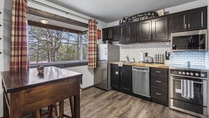 A modern kitchen with dark cabinets, a stainless steel refrigerator, a dishwasher, and an oven. There is a window with plaid curtains and a view of trees outside. - Charming Rustic Retreat - Hot Tub - Pool - Coffee - Fire Pit - 1 mile from SDC! (Branson)