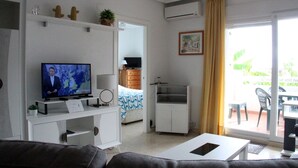 A living room with a television, a sofa, a dining table, and a balcony with a view of greenery.