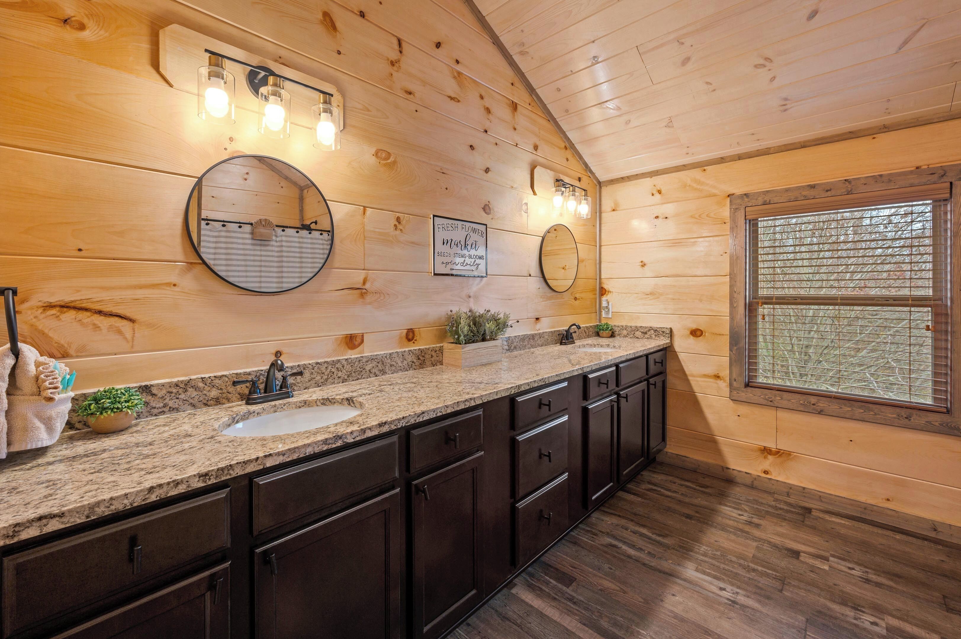 A wooden cabin bathroom with a granite countertop, two sinks, dark cabinets, and a large window with a view.