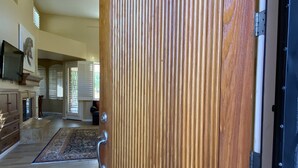 A wooden door with vertical grooves, a tiled floor, and a hallway leading to a living area with a fireplace and a television.