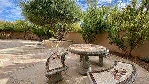 A patio area with a mosaic-tiled table and matching benches, surrounded by trees and a stone wall.