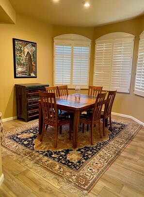 Comedor con mesa y sillas de madera, rodeado por un tapete estampado, con un cuadro en la pared y una ventana con persianas.