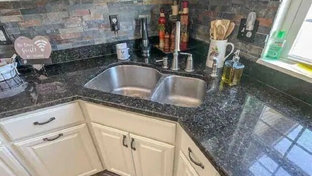 Een keuken met een granieten aanrecht, een dubbele spoelbak, witte kasten en een spatwand met kleurrijke tegels.