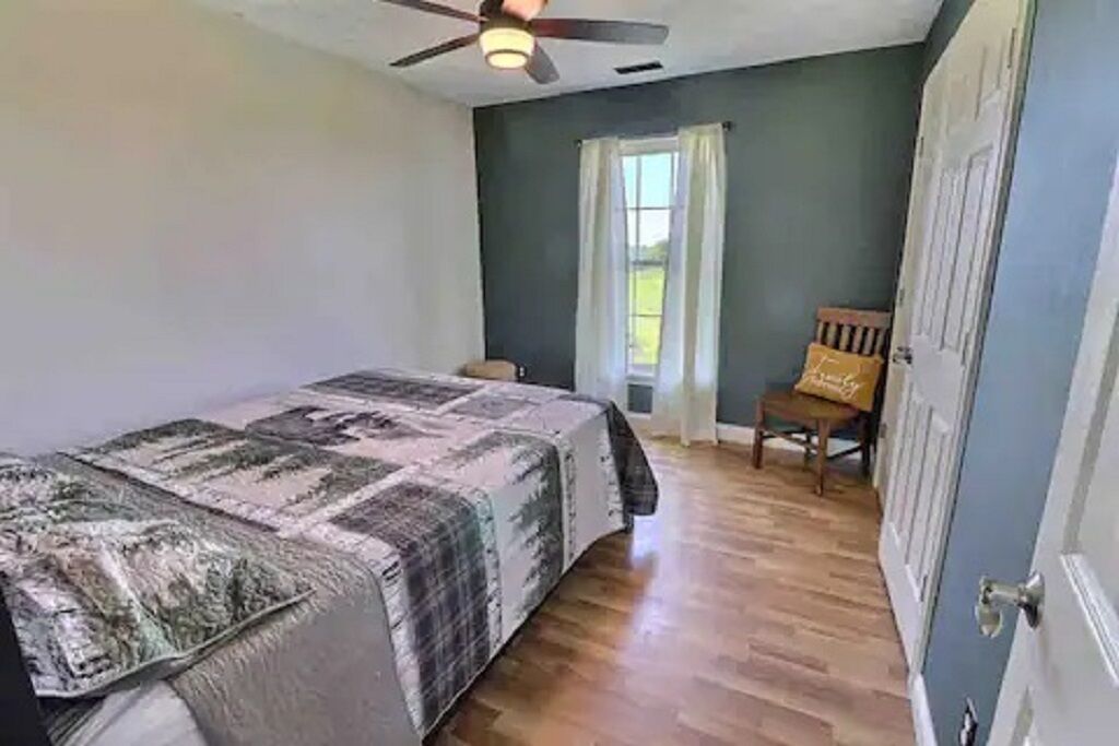 Um quarto com uma cama, um ventilador de teto, uma janela com cortinas e uma porta.