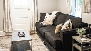 Un soggiorno con un divano grigio scuro, un tavolino da caffè e un comodino con una lampada. Si vede una porta con finestra e tende, e un pavimento in legno.