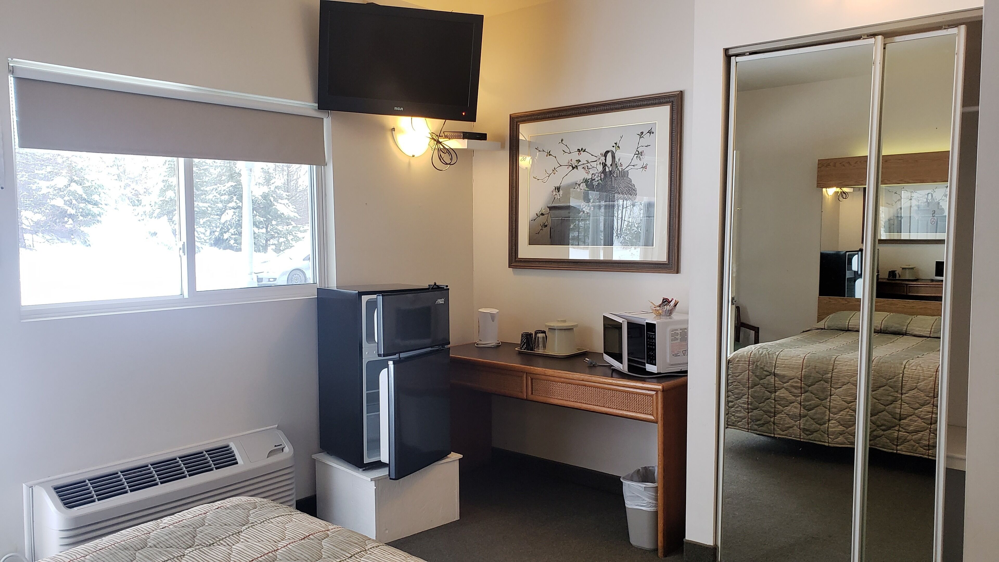 A compact hotel room with a kitchenette, a flat-screen TV, a framed artwork, and a large mirror.