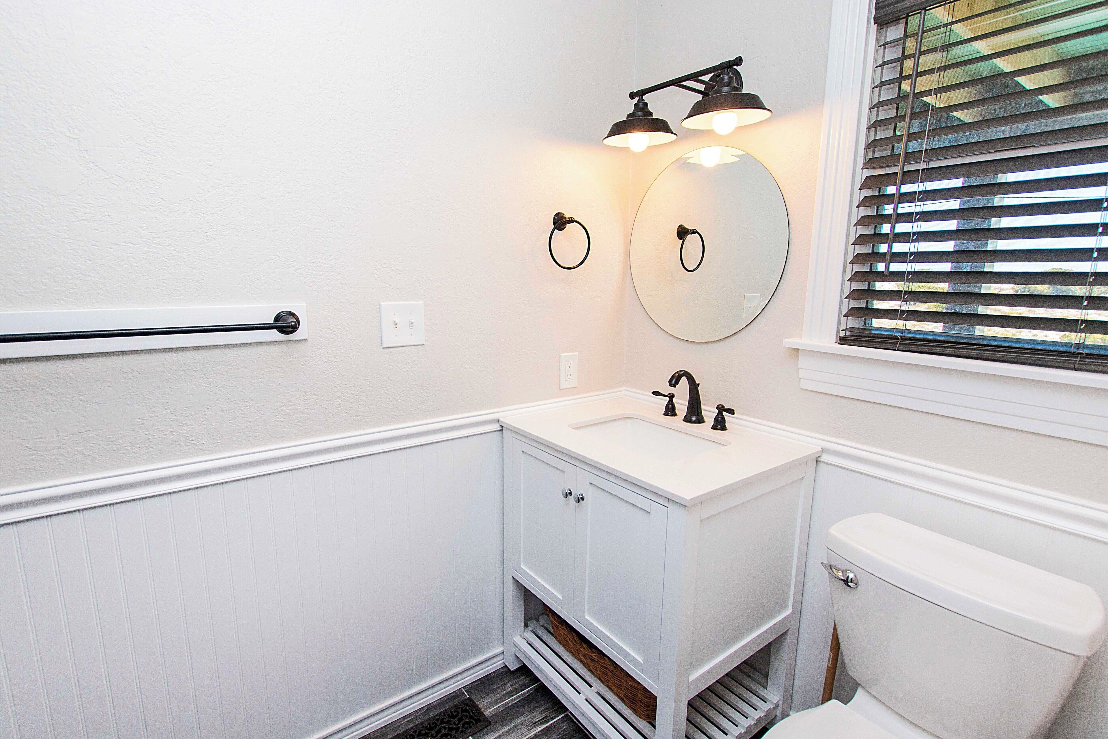 A bathroom with a white vanity, a round mirror, a toilet, and a window with blinds.