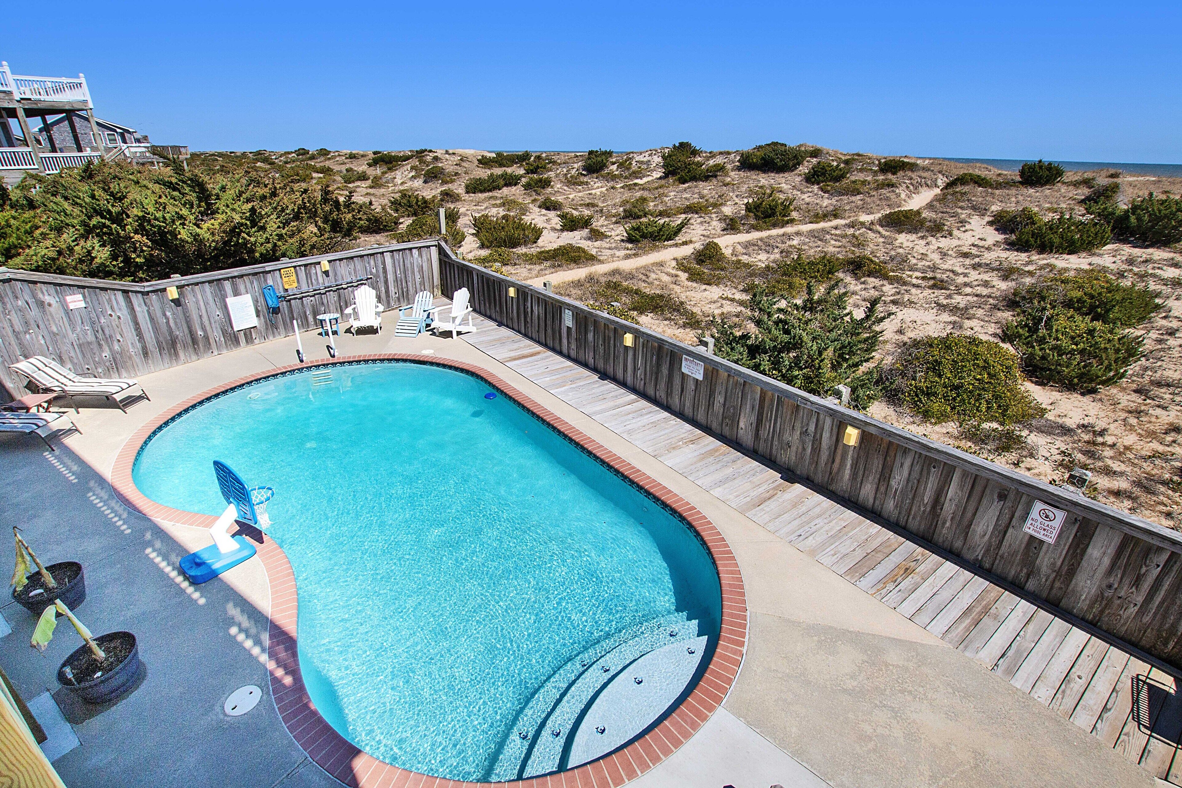 A swimming pool with a wooden deck, lounge chairs, and a view of the beach.