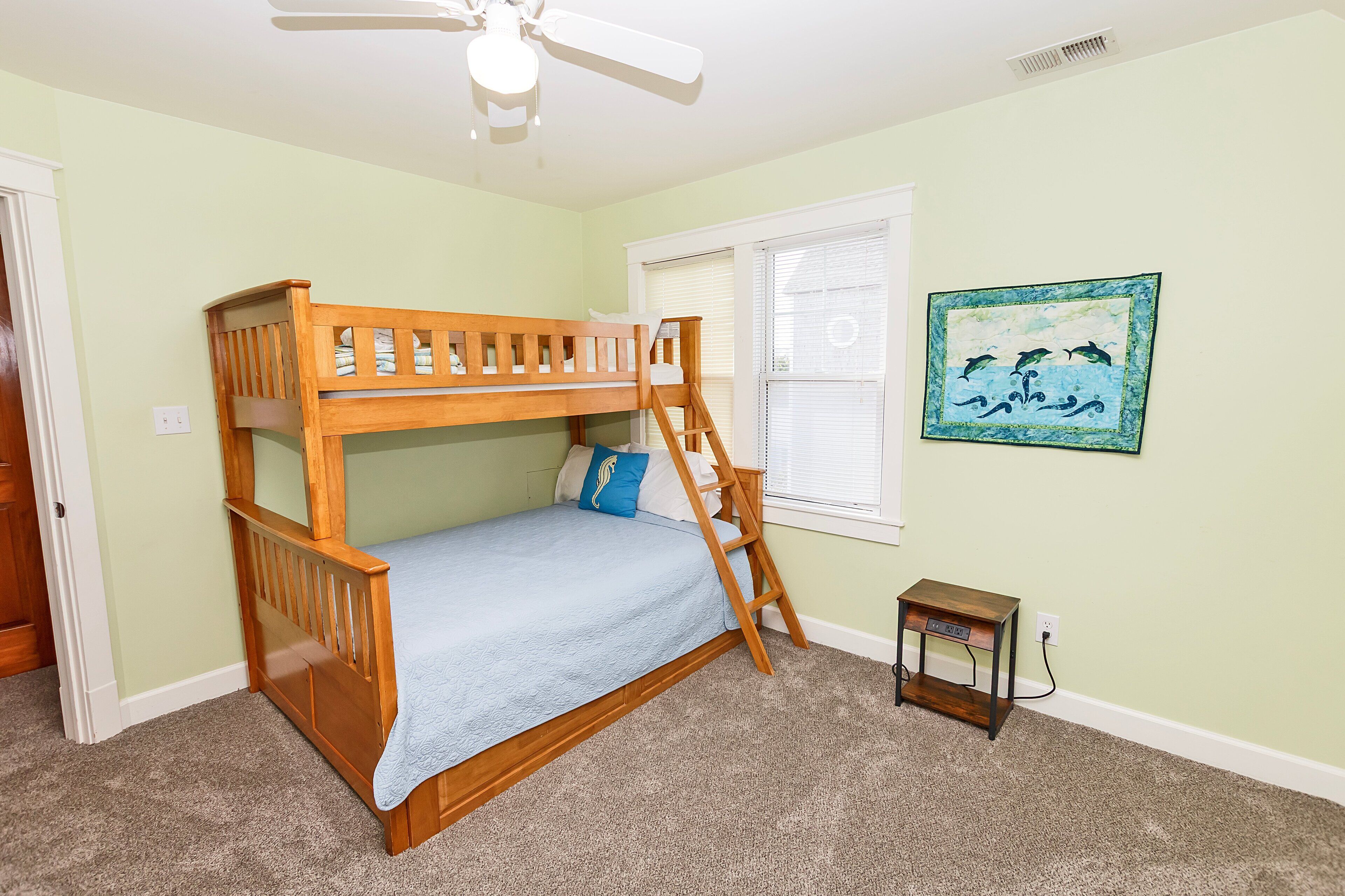 Une chambre avec un lit superposé, un ventilateur de plafond, une fenêtre avec des stores et une petite table.