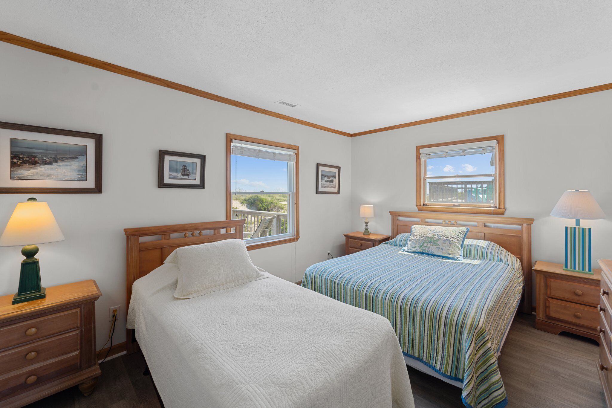 A bedroom with two beds, wooden headboards, bedside tables with lamps, framed pictures on the walls, and large windows offering a view of the outdoors.