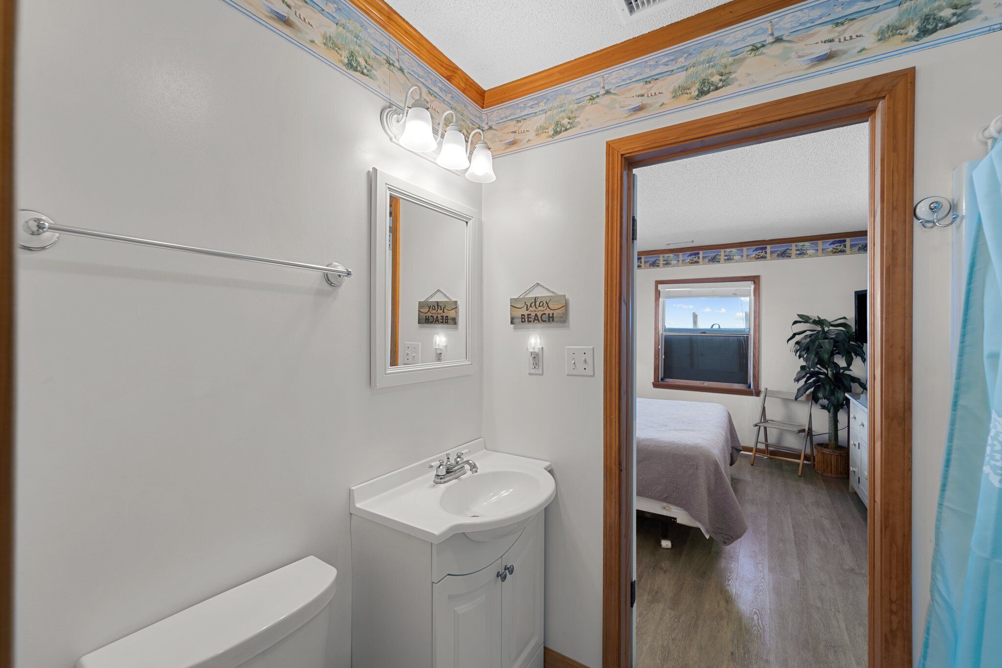 Une salle de bain avec un lavabo blanc, un miroir et un porte-serviettes. Une porte en bois donne sur une chambre à coucher équipée d’un lit et d’une fenêtre.